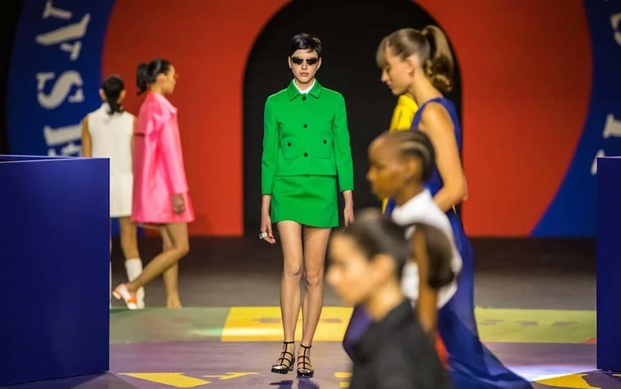 Kermit-green two-piece on the runway, with other models in pink and navy walking behind